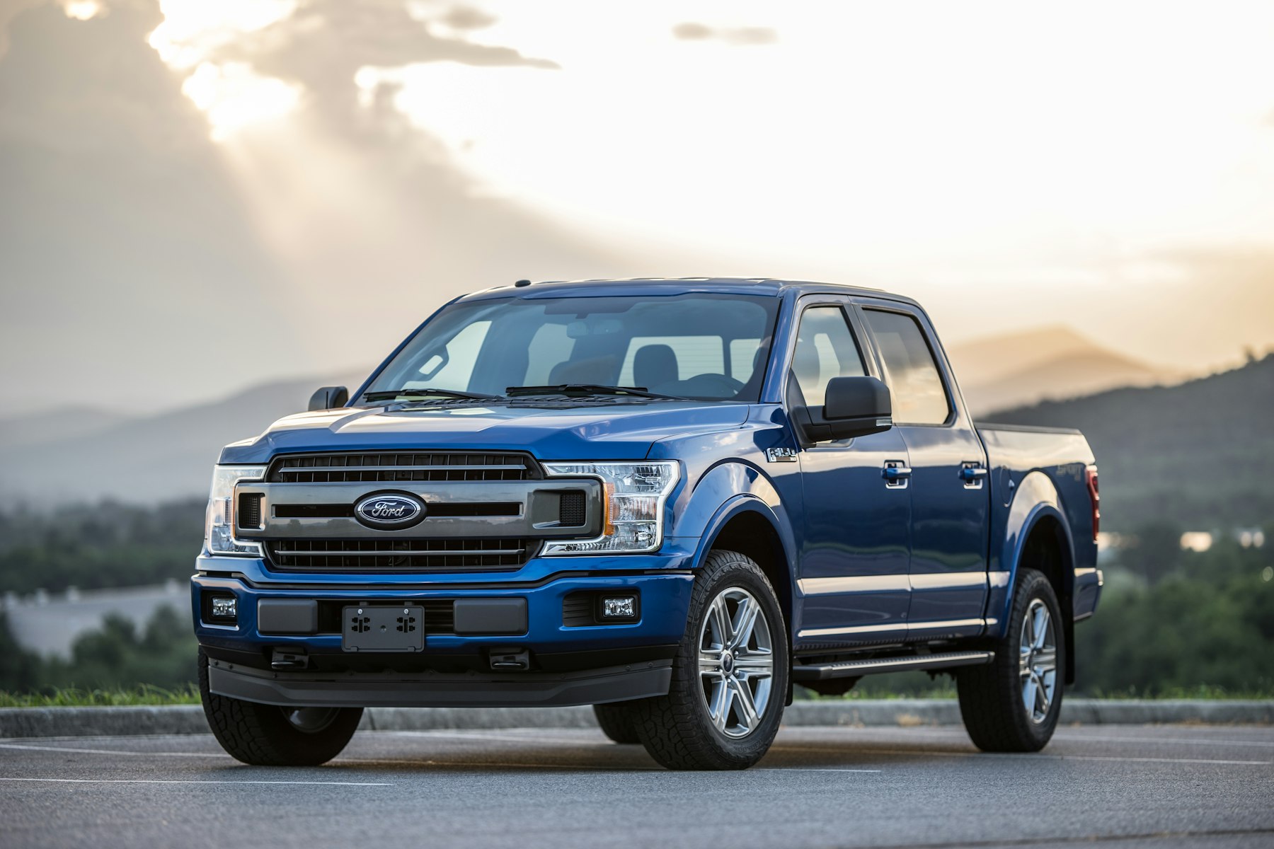 A blue crew-cab pickup truck parked on a ridge at sunset. Illustrative photography — not a vehicle held, offered for sale, or priced by Automotive Car Brokers.